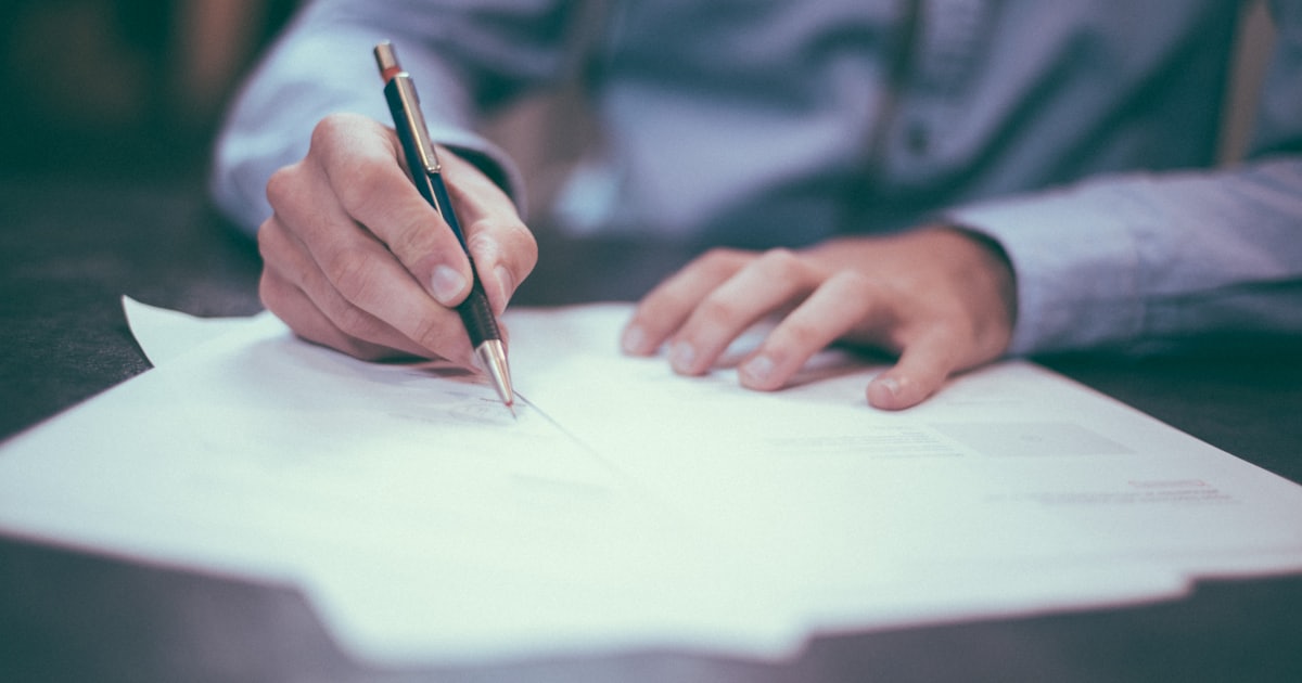 Mão assinando um documento com caneta, representando um contrato de sociedade