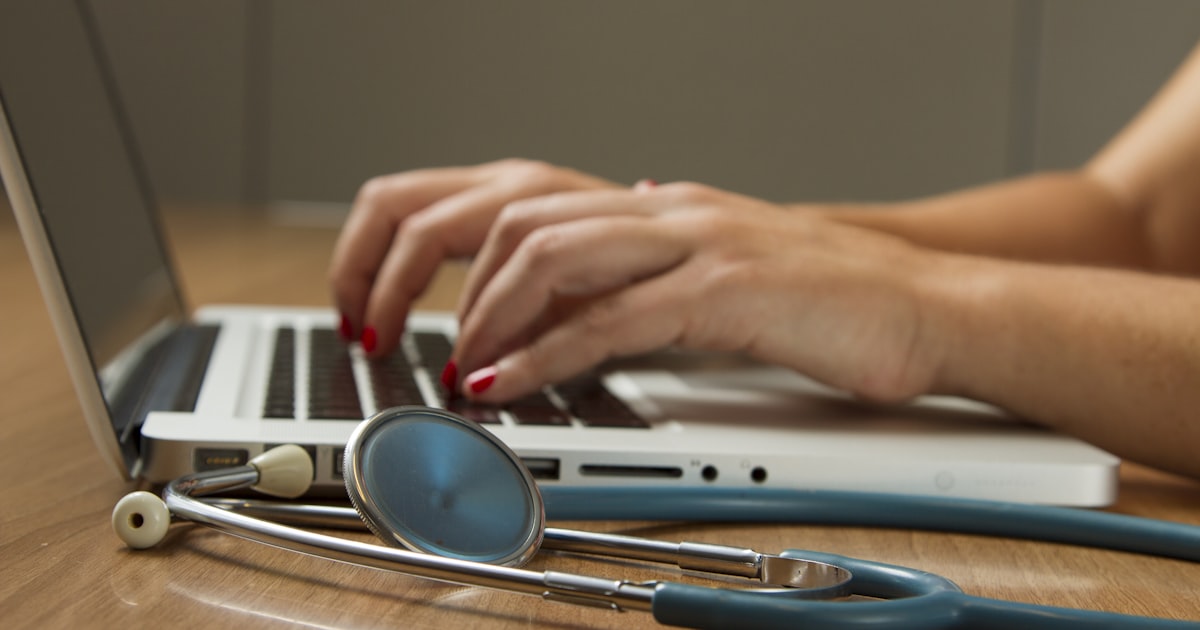 Mãos digitando em um laptop ao lado de um estetoscópio, representando o prontuário eletrônico